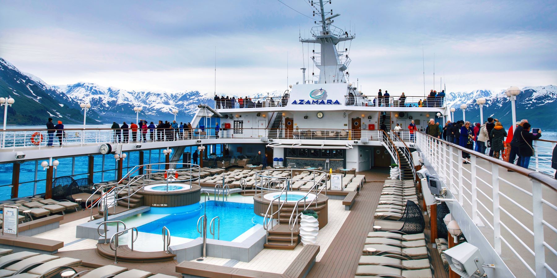 Luxury Azamara cruise ship sailing in Alaska, with guests enjoying panoramic views of snow-capped mountains and glaciers from the pool deck.