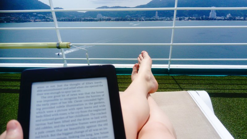 Woman sits on sun deck of cruise ship relaxing and reading her E-reader as the ship sails along through calm peaceful waters